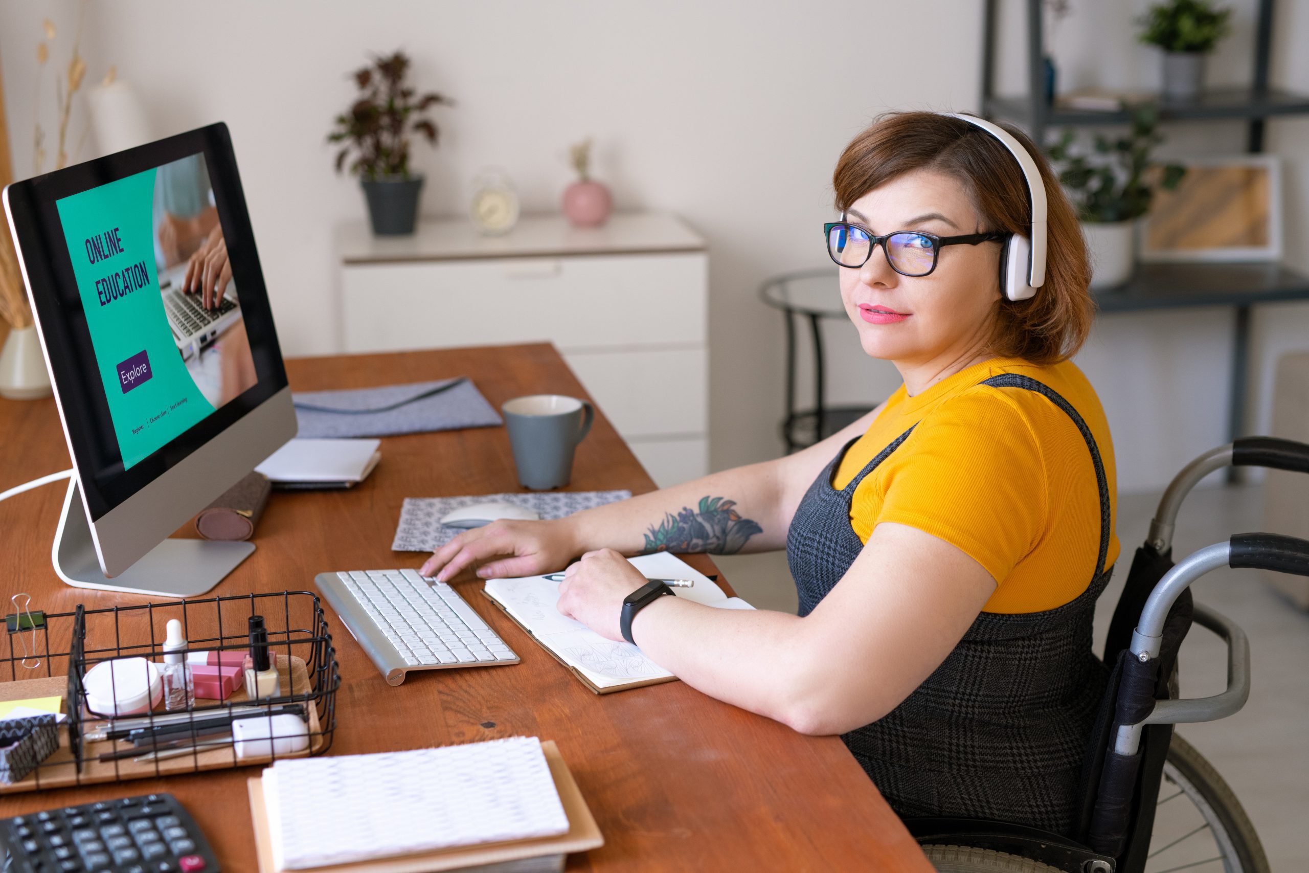Portrait of content young portly woman in wireless headphones sitting in wheelchair and using computer while enjoying online education